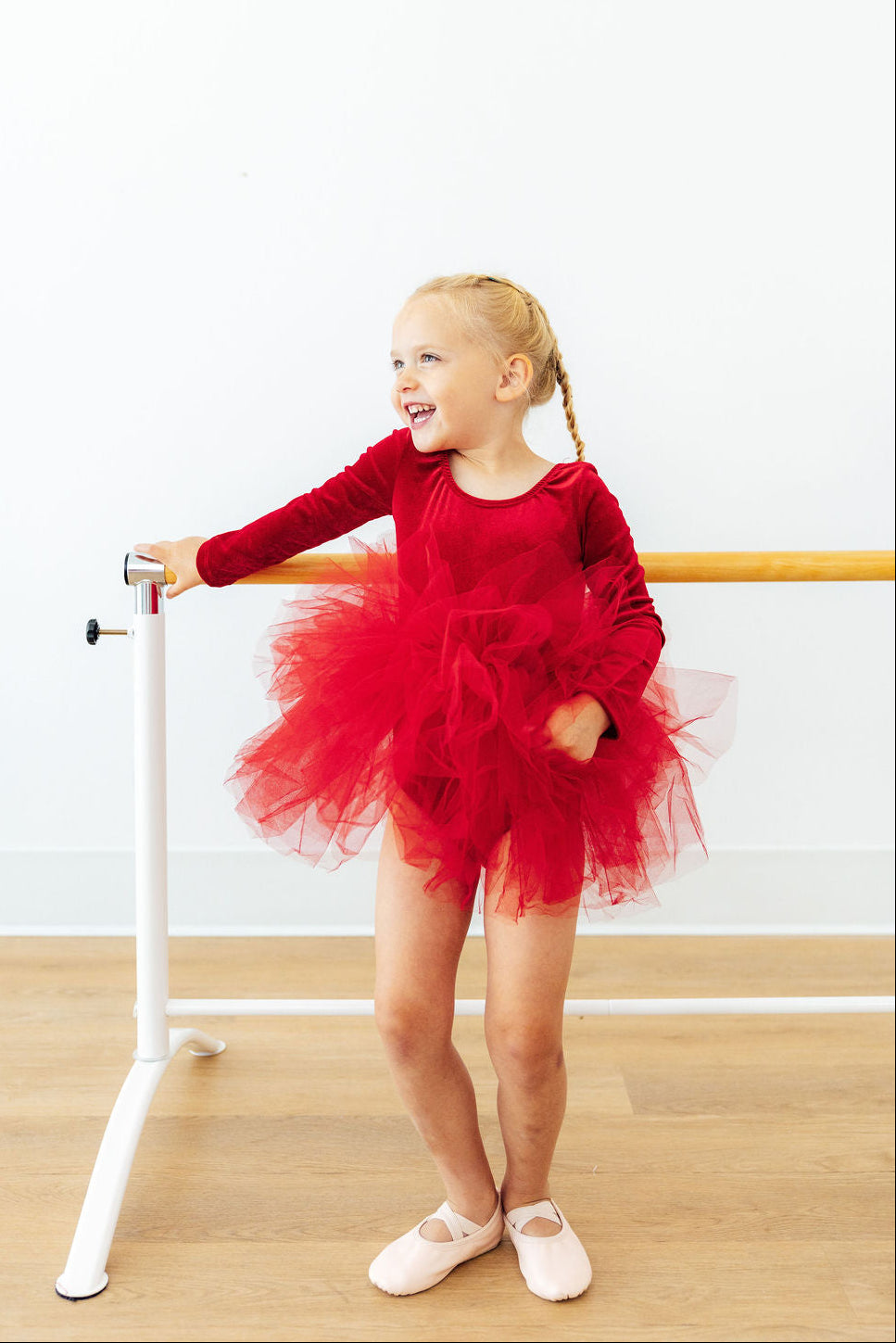 A young blonde girl in a red long-sleeve tutu dance dress and white ballet slippers poses next to a white ballet barre in a studio with a light wood floor.