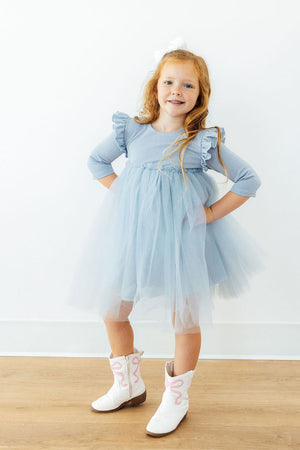 A young girl with red hair stands with hands on hips wearing a light blue tulle dress with ruffled sleeves and white cowboy boots, posing in front of a white wall and light wood floor.